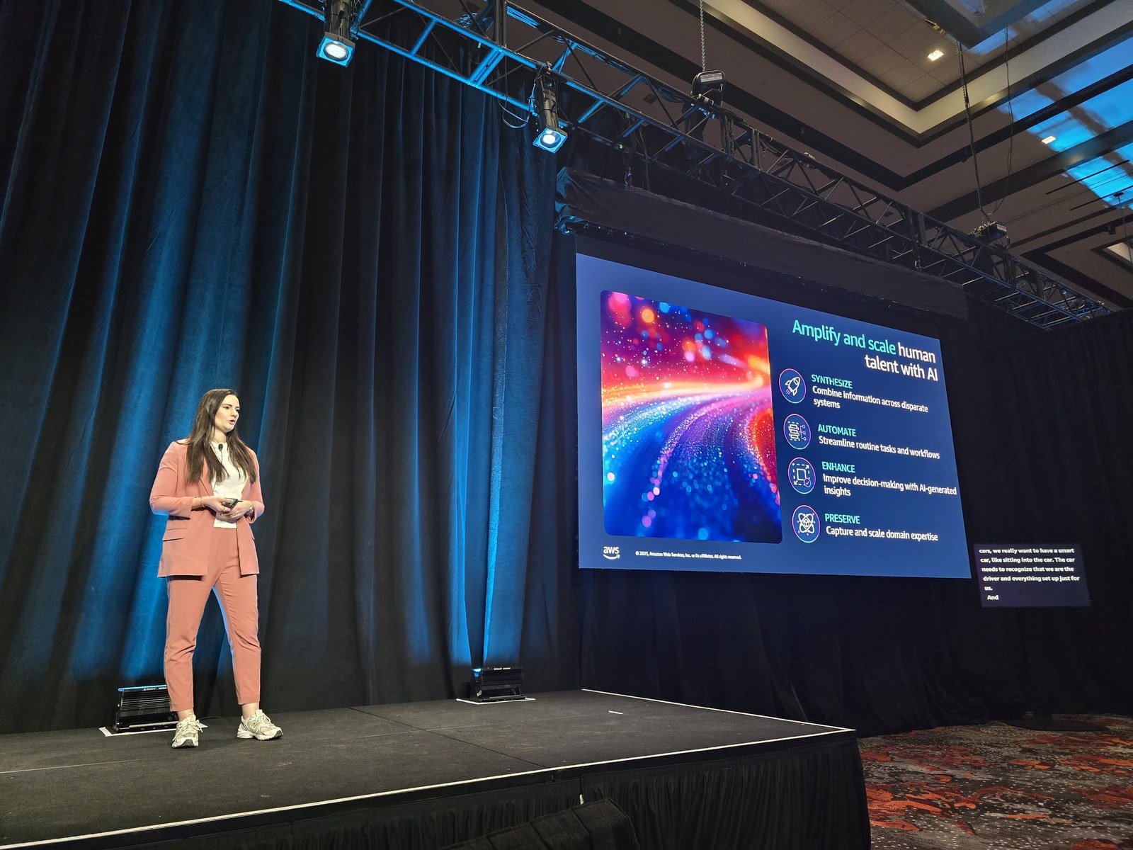 IND367 セッションのステージ。左に立つ Verena Koutsovagelis 氏、右手のスクリーンに「Amplify and scale human talent with AI」と題したスライドが表示され、SYNTHESIZE / AUTOMATE / ENHANCE / PRESERVE の 4 項目が並んでいる